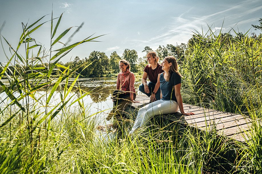 Drei Frauen sitzen auf einem Holzsteg über einem See