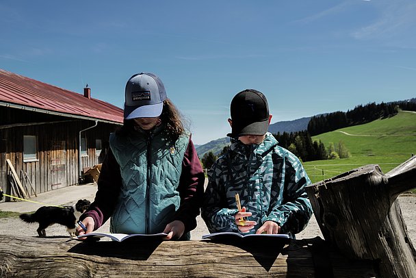 Junge und Mädchen mit Rätselheften vor einer Berghütte