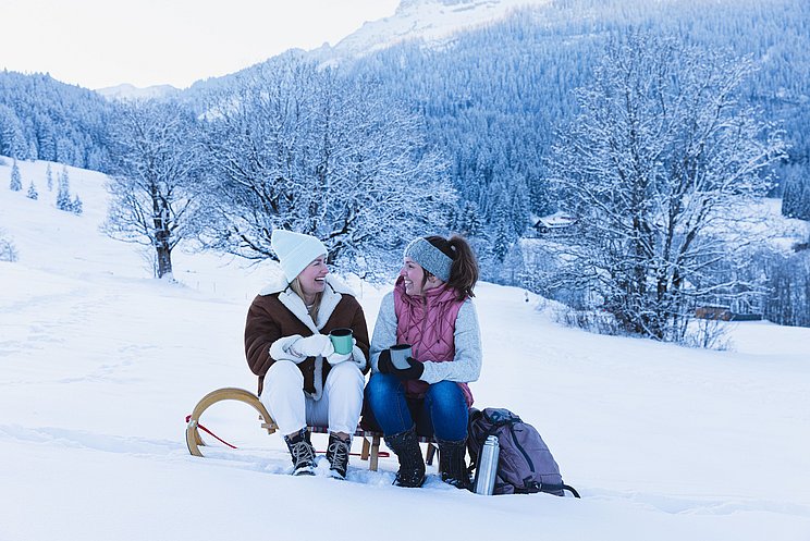 Zwei Freundinnen sitzen in einer Winterlandschaft auf Schlitten und trinken Tee