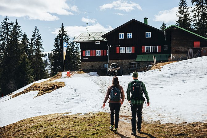 Mann und Frau wandern auf das Berghaus Schwaben zu, es ist Spätwinter, es ist sonnig und liegt nur noch teilweise Schnee