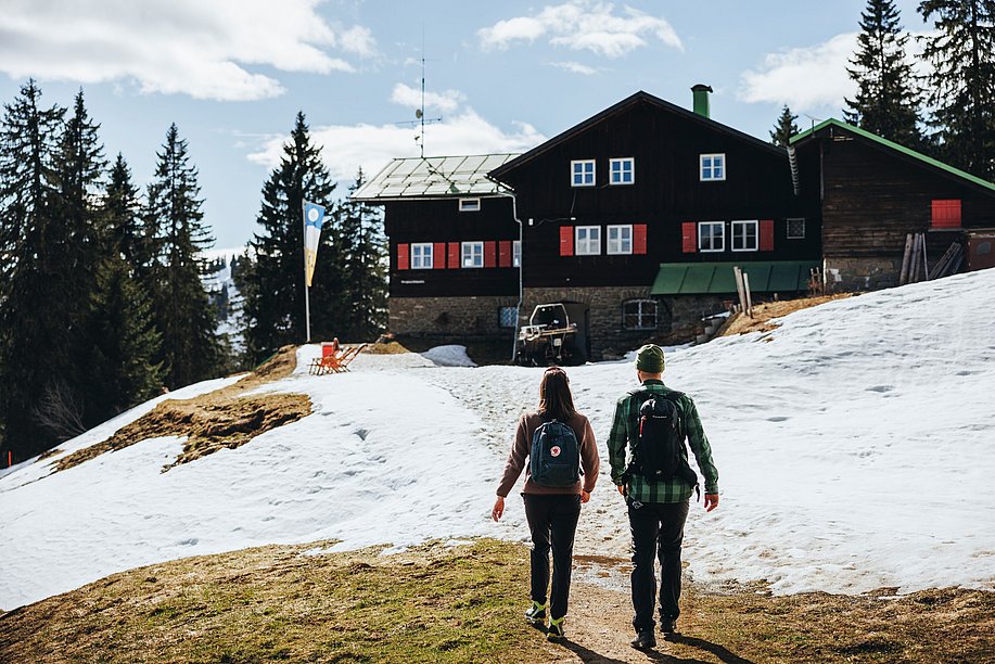 Mann und Frau wandern auf das Berghaus Schwaben zu, es ist Spätwinter, es ist sonnig und liegt nur noch teilweise Schnee Mann und Frau wandern auf das Berghaus Schwaben zu, es ist Spätwinter, es ist sonnig und liegt nur noch teilweise Schnee