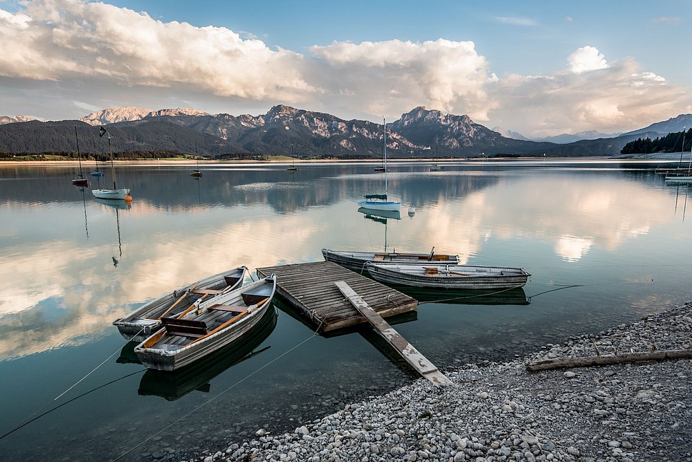 Ruderboote liegen an einer schwimmenden Plattform im Forggensee, im Hintergrund die Berge