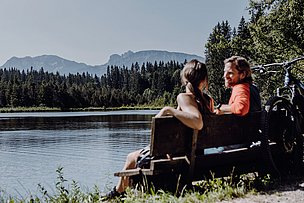 Paar sitzt auf einer Bank am Kögelweiher, daran lehnt ein Fahrrad, im Hintergrund die Alpen