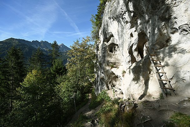 Felswand mit Höhlen, Holztreppe in Höhle, Bank und Bergblick