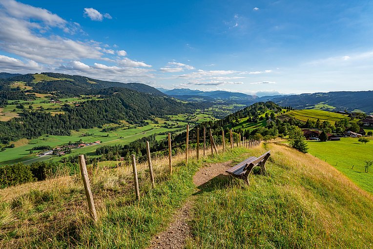 Bank an einem Wanderweg mit Ausblick über Berge mit Wiesen und Wäldern