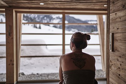 Frau von hinten guckt durch ein Panoramafenster aus der Sauna hinaus in die Schneelandschaft