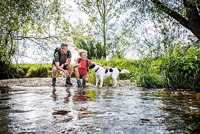 Mann und Frau in Wanderklamotten mit Hund an einem Flussufer