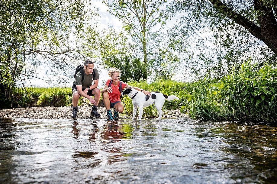 Mann und Frau in Wanderklamotten mit Hund an einem Flussufer