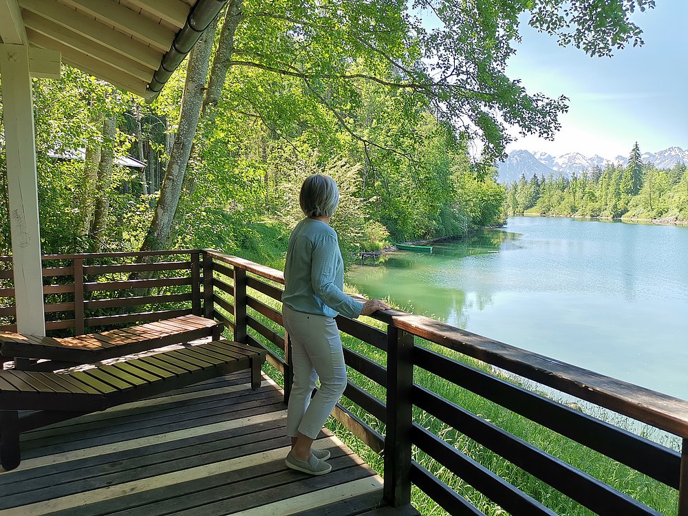 Frau auf einer Holzterrasee blickt über den Auwaldsee in die Berge