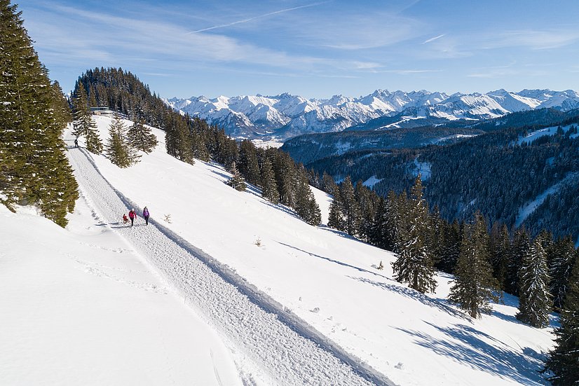 Winterwanderweg in der Höhe mit zwei Wandernden und einem Kind auf einem Schlitten mit Bergpanorama