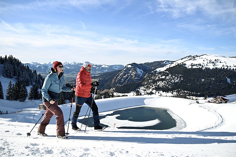 Mann und Frau beim Winterwandern auf präpariertem Weg, im Hintergrund ein Speicherteich, verschneite Berge und ein grünes Tal