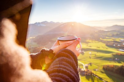 Frau hält eine Teetasse Richtung Sonne, im Hintergrund Tal und Berge