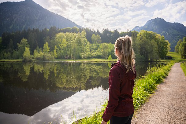 Frau steht am Moorweiher, die Berge spiegeln sich im Wasser