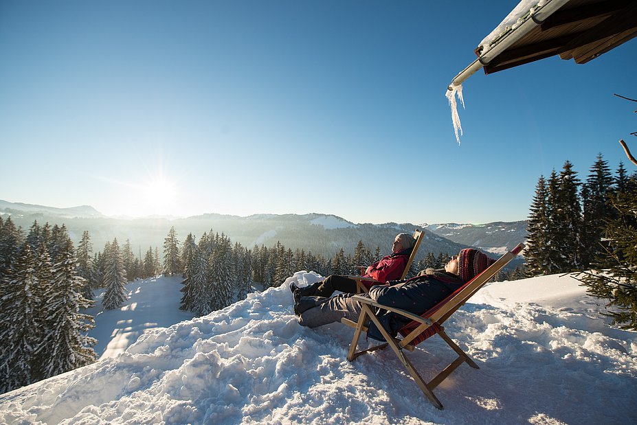 Zwei Winterwanderer entspannen in Liegestühlen in der Sonne an einer Hütte