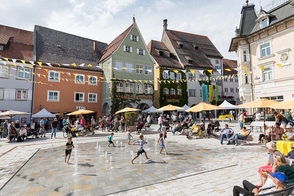 Kinder toben in Wasserfontänen auf Marktplatz in Isny, Menschen sitzen unter bunten Sonnenschirmen