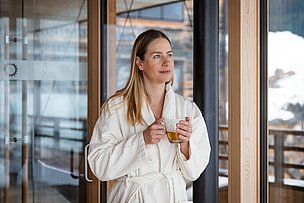 Frau im Bademantel steht mit Tee in der Hand am Fenster