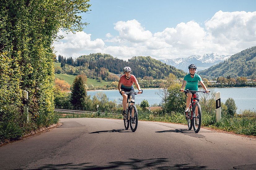 Zwei Frauen fahren Fahrrad rund um den Großen Alpsee. Hinter ihnen der Große Alpsee mit tollem Bergpanorama.