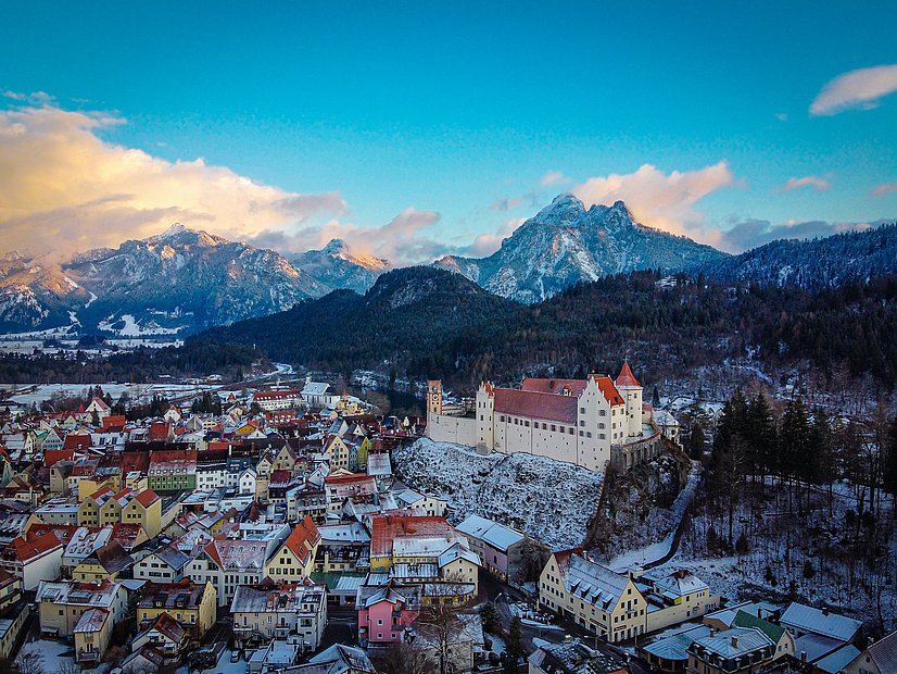 Luftansicht vom mit Schnee bepuderten Füssen, prominent das Hohe Schloss und im Hintergrund die Berge
