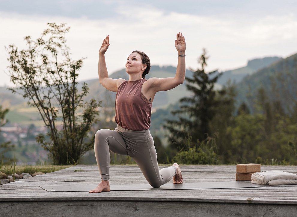 Frau macht Yoga auf einer Holzplattform, im Hintergrund die Berge