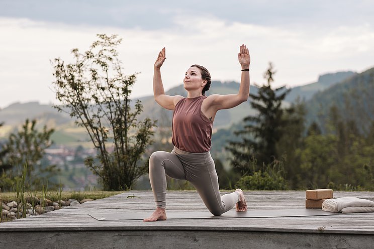 Frau macht Yoga auf einer Holzplattform, im Hintergrund die Berge
