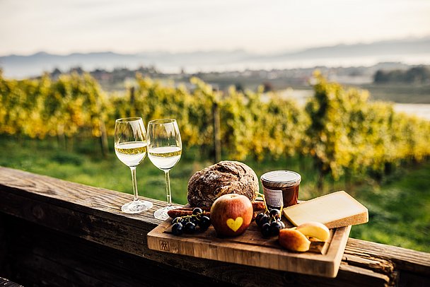 Eine Brotzeit und gefüllte Weißweingläser, im Hintergrund ist der Bodensee zu sehen