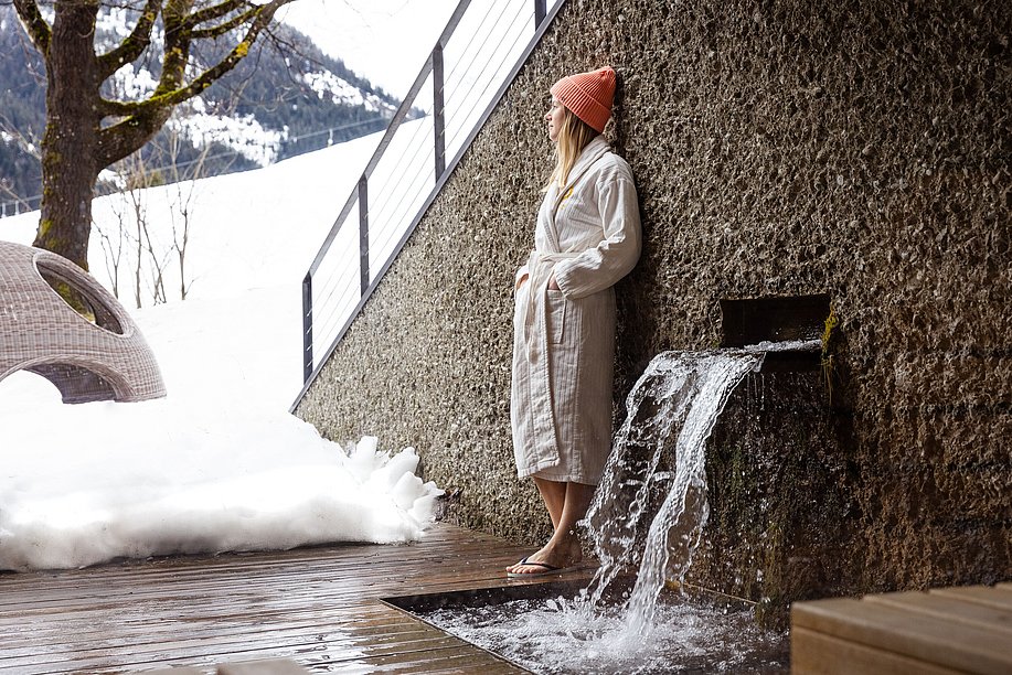 Frau steht in Bademantel und mit Mütze im Außenbereich vom Wellnessbereich, schaut über Schneelandschaft