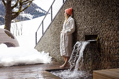 Frau steht in Bademantel und mit Mütze im Außenbereich vom Wellnessbereich, schaut über Schneelandschaft