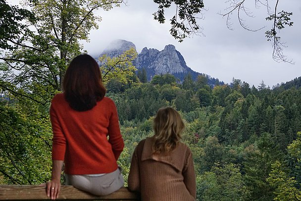 Zwei Frauen von hinten auf einer Bank gucken über Wald zu einem Gipfel