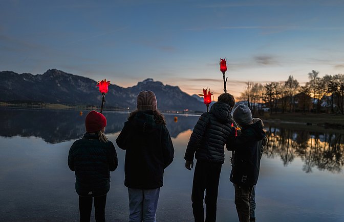 Vier Kinder von hinten am Forggensee mit selbstgebastelten, leuchtenden Fackeln in der Hand Vier Kinder von hinten am Forggensee mit selbstgebastelten, leuchtenden Fackeln in der Hand