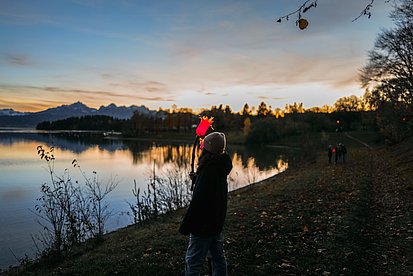 Ein Mädchen steht mit ihrer selbstgebastelten, leuchtenden Fackel an einem Seeufer, es dämmert