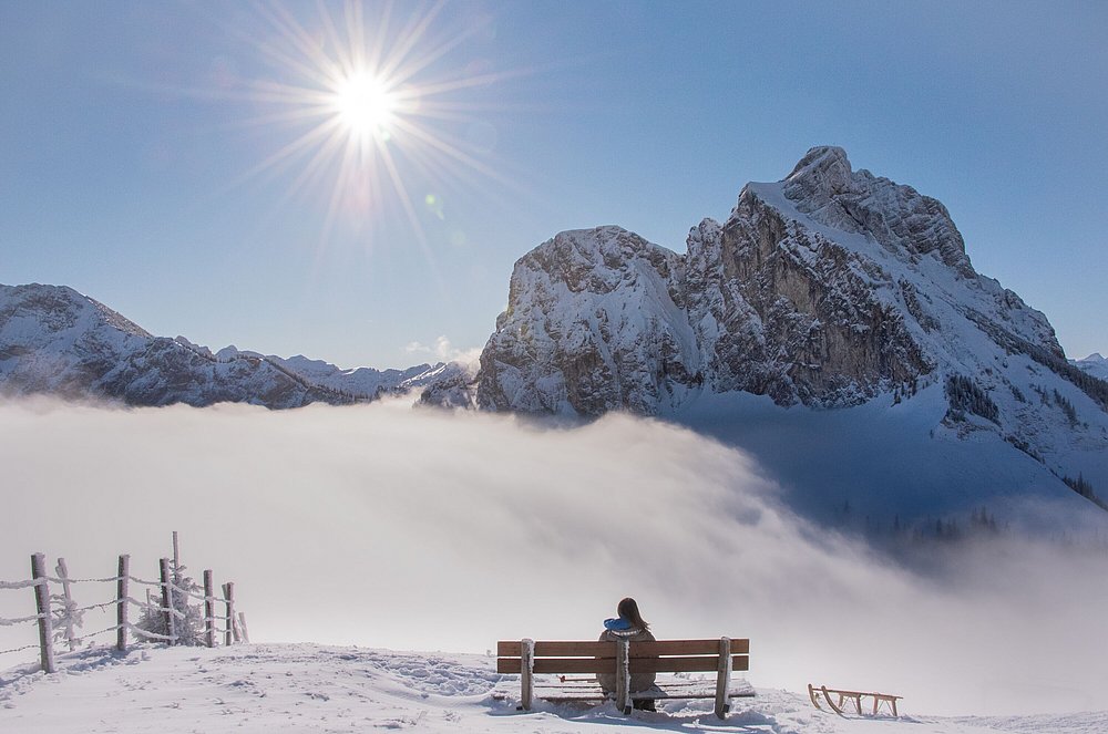 Eine Wanderin mit Schlitten sitzt auf einer Bank und schaut zum Aggenstein. Es liegt Schnee, die Sonne strahlt am blauen Himmel .