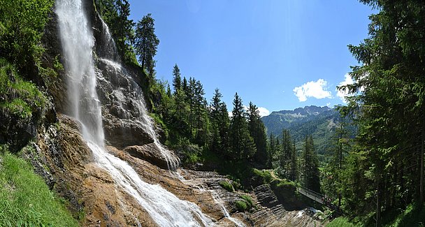 Wasserfall an Felswand, Brücke über Bach, im Hintergrund Berge
