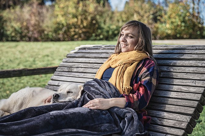 Frau sitzt zufrieden mit einem Hund und einer Decke über den Beinen auf einer Sonnenliege