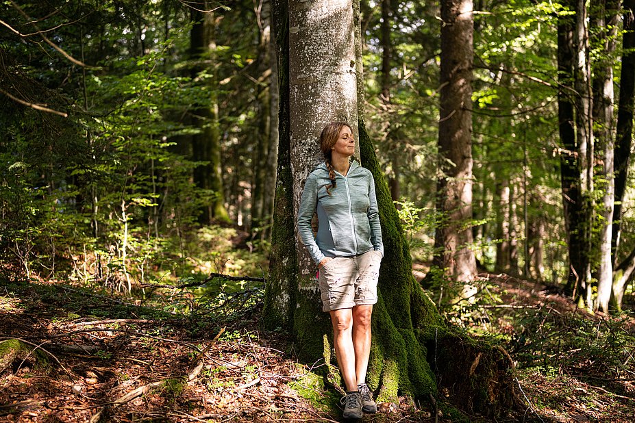 Eine Frau lehnt mit geschlossenen Augen im Wald an einem Baumstamm