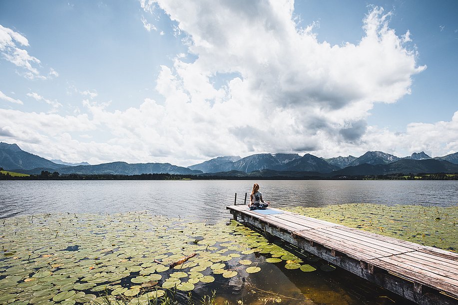 Frau sitzt im Schneidersitz auf einem Holzsteg am Hofpensee, im Hintergrund die Alpenkette