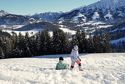 Zwei Kinder spielen im Schnee. Im Hintergrund ist Bergpanorama und strahlend blauer Himmel. 