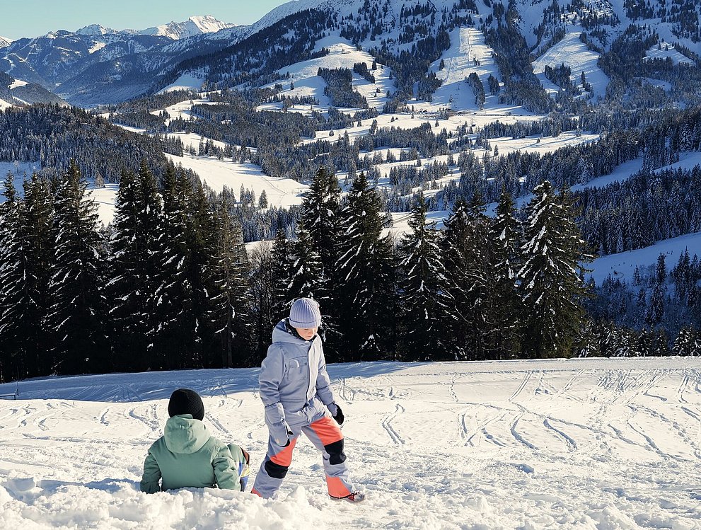 Zwei Kinder spielen im Schnee. Im Hintergrund ist Bergpanorama und strahlend blauer Himmel. 