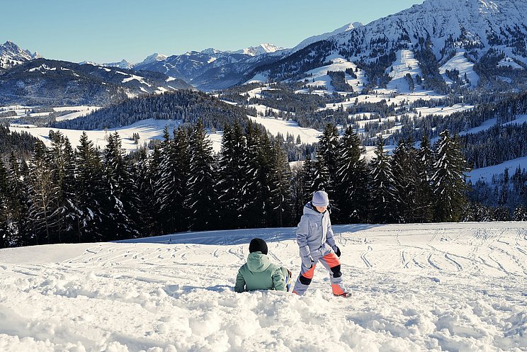 Zwei Kinder spielen im Schnee. Im Hintergrund ist Bergpanorama und strahlend blauer Himmel. 