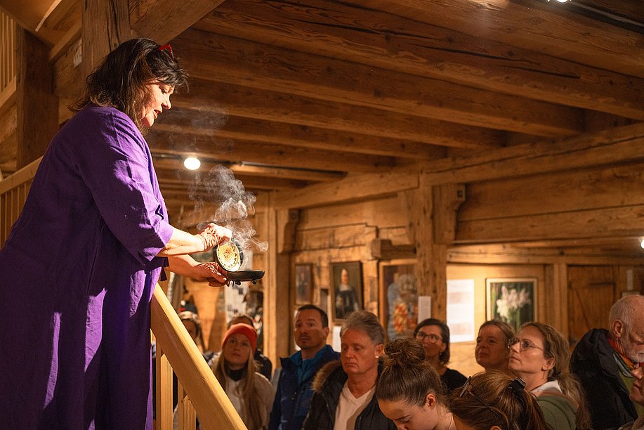 Eine Frau räuchert in einem Holzhaus vor Publikum