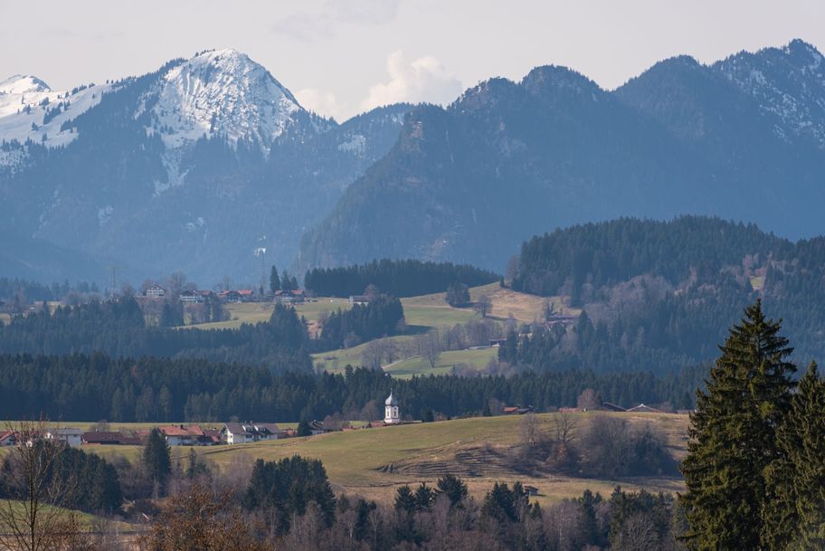 Der Kirchturm der St. Martinskirche Hopferua spitzelt hinter den Hügeln hervor. Im Hintergrund die imposanten Berge.